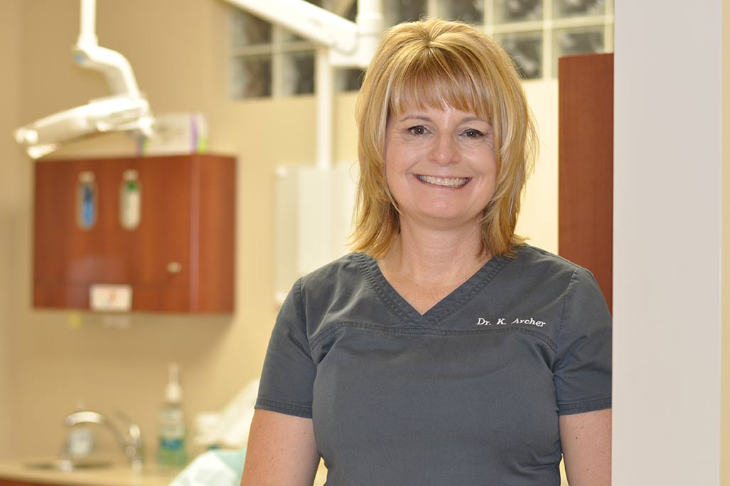 Dr. Karen Archer, DDS at Karen Archer DDS in Leamington, ON, wearing gray scrubs in front of a dental service room