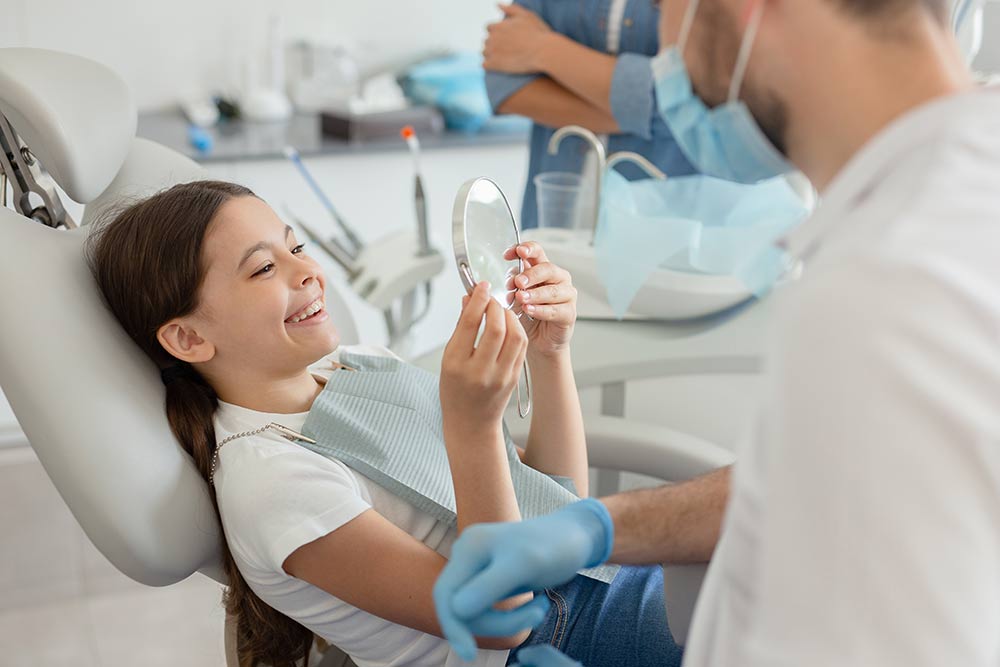 A young child smiles and looks into a mirror as she sits on a dental chair next to two other people