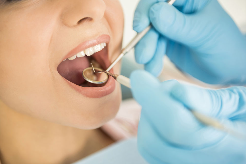 A dental patient opens their mouth as a dentist with blue rubber gloves uses a mirror and pick to clean the teeth